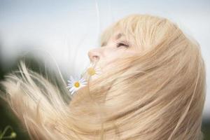 un mujer con fluido blanco pelo sostiene un manzanilla flor en su boca.hermosa rubia cabello, flores y verano. foto