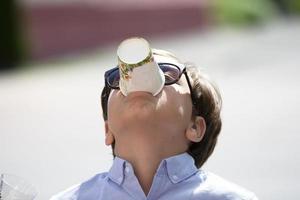 The child wants to drink in the heat. A boy in sunglasses holds a plastic cup in his mouth. photo