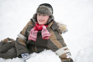 A boy sits in a snowdrift on a winter day and smiles. Child on a walk in winter. photo