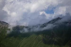 Dark atmospheric surreal landscape with a dark rocky mountain peak in low clouds in a gray cloudy sky. A gray low cloud on a high peak. High black rock in low clouds. Surreal gloomy mountains. photo