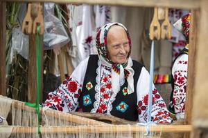 An old Belarusian or Ukrainian woman in an embroidered shirt at a vintage loom. Slavic elderly woman in national ethnic clothes. photo