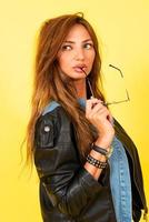 Yellow background, a girl in a leather jacket in the style of rock, shows emotions, holds glasses in her teeth photo