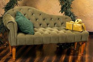 Christmas composition with decoration, christmas tree branch, stylish green classic sofa, gift boxes and accessories in cozy home decor. Happy xmas and New Year holiday concept. Vertical photo