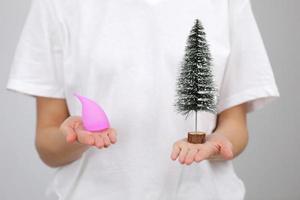 Close up of a young woman hands holding reusable pink silicone menstrual cup and Christmas tree on white t-shirt background. Zero waste menstruation hygiene concept. photo