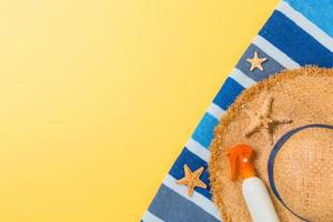 Beach flat lay accessories with copy space. Striped blue and white towel, seashells, staw sunhat and a bottle of sunblock on yellow background. Summer holiday concept photo