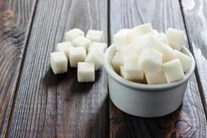 azúcar blanco en un tazón sobre fondo de madera. enfoque selectivo, horizontal. unos terrones de azúcar están cerca del vaso lleno de azúcar blanca. consumo de malas calorias foto