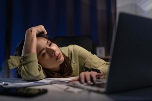 soñoliento agotado asiático joven mujer de negocios trabajando a oficina escritorio con su computadora portátil, su ojos son clausura. tiempo extraordinario trabajando concepto foto