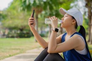 Young beautiful asian drinking water from bottle and using smartphone, she resting after jogging at green park photo
