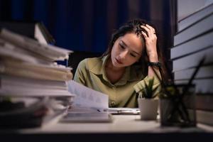 Asian businesswoman working hard late hours with a lot of document at home. She is busy and exhausted of work overtime at night. photo