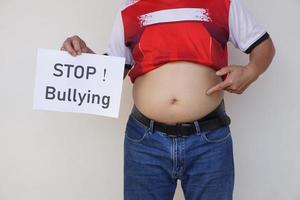 Closeup man with his big belly holds paper sign with word STOP  Bullying .Concept, campaign for anti bullying, body shaming or parody of others physical appearance. photo