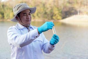 El investigador ambiental del hombre asiático sostiene un tubo de agua de muestra para inspeccionar desde el lago. concepto, exploración, análisis de la calidad del agua de fuente natural. investigación de campo en ecología. foto