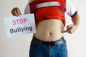 Closeup man with his big belly holds paper sign with word  STOP  Bullying . Concept, campaign for anti bullying, body shaming or parody of others physical appearance. photo