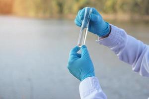 Closeup researcher hands wears blue gloves holds test glass tube that contain sample water from the lake. Concept, explore, analysis water quality from natural source. Ecology field research. photo