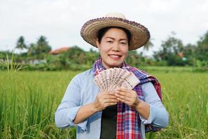 asiático granjero mujer es a arrozal campo, usa sombrero y rojo tartán camisa, sostener tailandés billete de banco dinero. concepto , granjero contento a obtener ganancia, ingreso, agricultura secundario dinero. foto