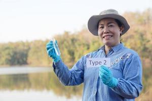 Asian woman ecologist hold tube of sample water and tag with word Type 2 for inspect and research water quality at the lake. Concept, explore, analysis water quality from natural source. Ecology field photo