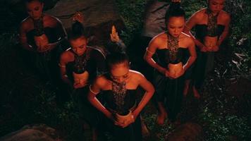 a group of Asian women standing together in front of big rocks while holding bamboo masks in their hands with flat expressions in the middle of the forest video