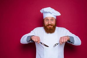 temeroso cocinero con barba y rojo delantal sostiene cuchillería en mano foto