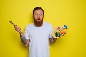 Undecided man with beard and tattoo is ready to draw with brushes photo