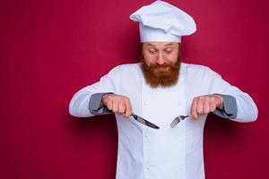 escéptico cocinero con barba y rojo delantal sostiene cuchillería en mano foto