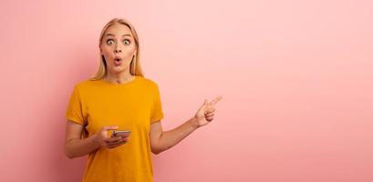 Blonde cute girl, with her smartphone, indicates something. Amazed and surprised expression face. Pink background with blank space for your text photo