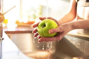 Young woman washes, with running water, an apple in the kitchen sink illuminated by the sun photo