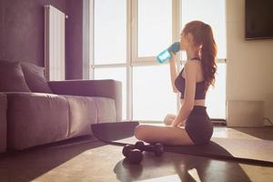 Young woman drinks water from the bottle after doing physical training exercises at home on the mat photo
