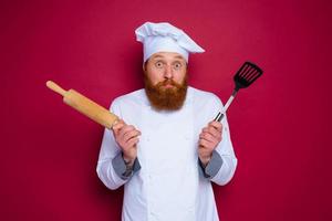 escéptico cocinero con barba y rojo delantal cocinero sostiene de madera laminación alfiler foto