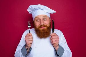 contento cocinero con barba y rojo delantal sostiene cuchillería en mano foto
