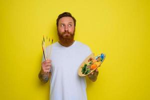 Undecided man with beard and tattoo is ready to draw with brushes photo