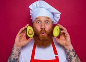 amazed chef with beard and red apron holds an avocado photo