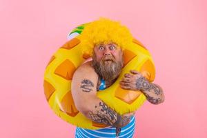 grasa asombrado hombre con peluca en cabeza es Listo a nadar con un rosquilla salvador de la vida foto