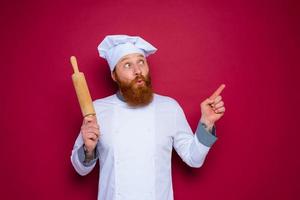 amazed chef with beard and red apron chef holds wooden rolling pin photo