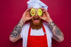 happy chef with beard and red apron holds an avocado photo