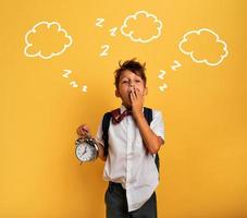 Young child student with ringing alarm clock is lazy going to school. Yellow background. photo