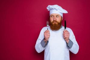 pensive chef with beard and red apron holds cutlery in hand photo