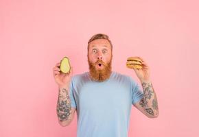 Amazed man with beard and tattoos is undecided if to eat an avocado or an hamburger photo