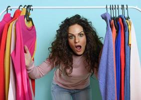 Woman with surprised expression chooses the clothes to buy in a store. Concept of shopping and shopaholic.Cyan background photo