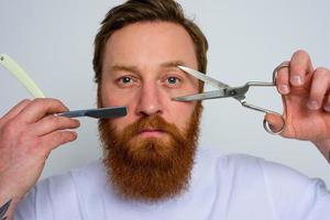 Worried man with scissors and blade is ready to cut the beard photo