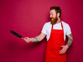 Happy chef with beard and red apron cooks with pan photo