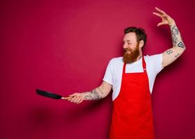 Happy chef with beard and red apron cooks with pan photo