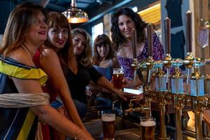 grupo de sonriente maduro mujer es Bebiendo cerveza en un pub en frente de un cerveza grifo foto