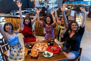 grupo de maduro mujer sentado en un pub aumento su manos mientras comiendo Pizza foto