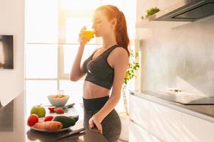 Athletic young red haired woman in the kitchen drinks a glass of fruit centrifuged juice photo