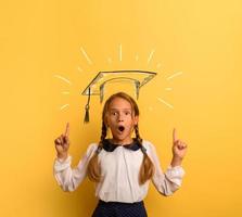 Young student is with shocked expression and indicates a graduation hat. Yellow background photo