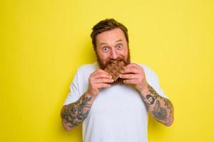 Happy man with beard and tattoos eats chocolate photo