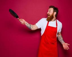 Happy chef with beard and red apron cooks with pan photo