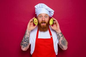 afraid chef with beard and red apron holds an avocado photo