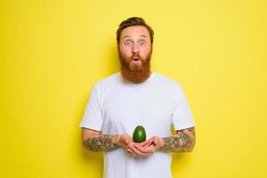 Amazed with beard and tattoos holds an avocado photo