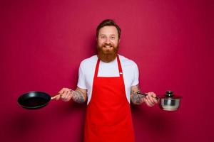 Happy chef with beard and red apron cooks with pan and pot photo