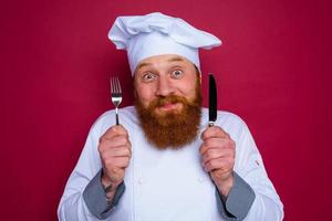 happy chef with beard and red apron holds cutlery in hand photo